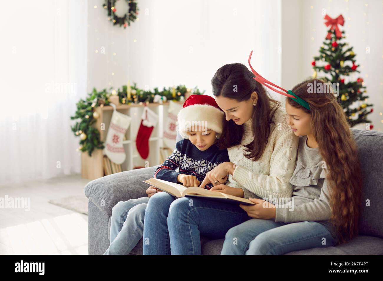 Madre amorevole con i suoi figli leggere un libro di storie in un accogliente e luminoso soggiorno il giorno invernale innevato. Foto Stock