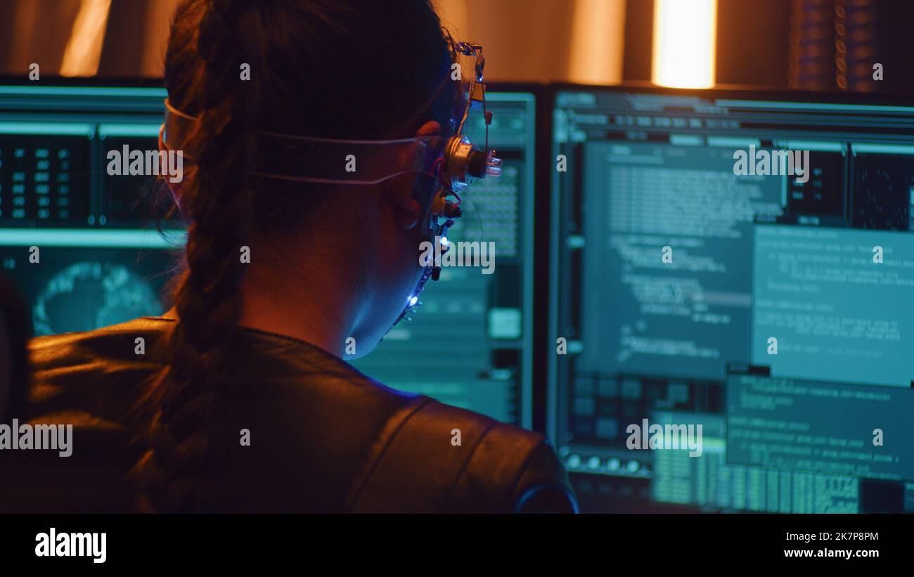 La ragazza funziona con schermo multiplo del calcolatore. Indossare un auricolare con LED che si illuminano durante la programmazione sul computer. Codifica. Ragazza dai capelli intrecciati in stile cyberpunk. Foto Stock