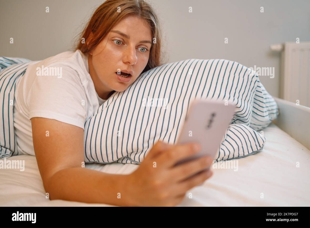 Scioccato giovane donna che si posa a letto e guardando la sveglia a smartphone in camera da letto a casa. Foto Stock