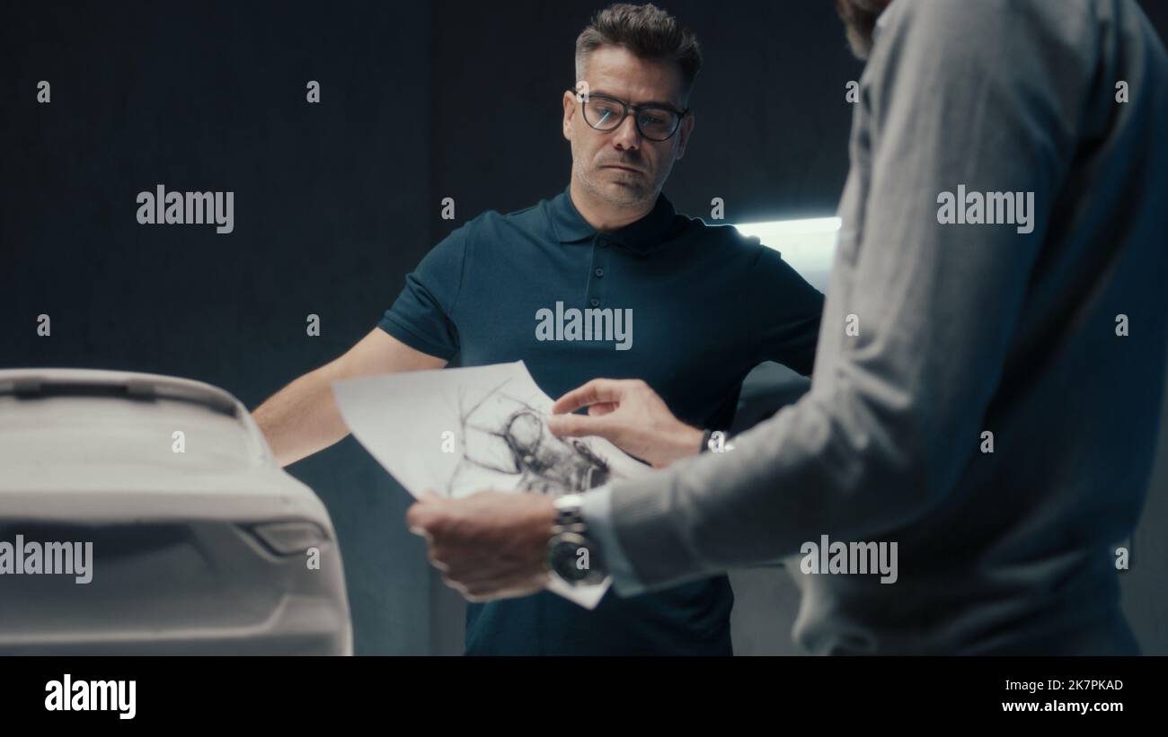 Progettisti e sviluppatori di auto esperti discutono del design della vettura mentre si trovano di fronte alla scultura della vettura prototipo. Utilizzando gli schizzi a matita in un'azienda di produzione di automobili ad alta tecnologia. Foto Stock