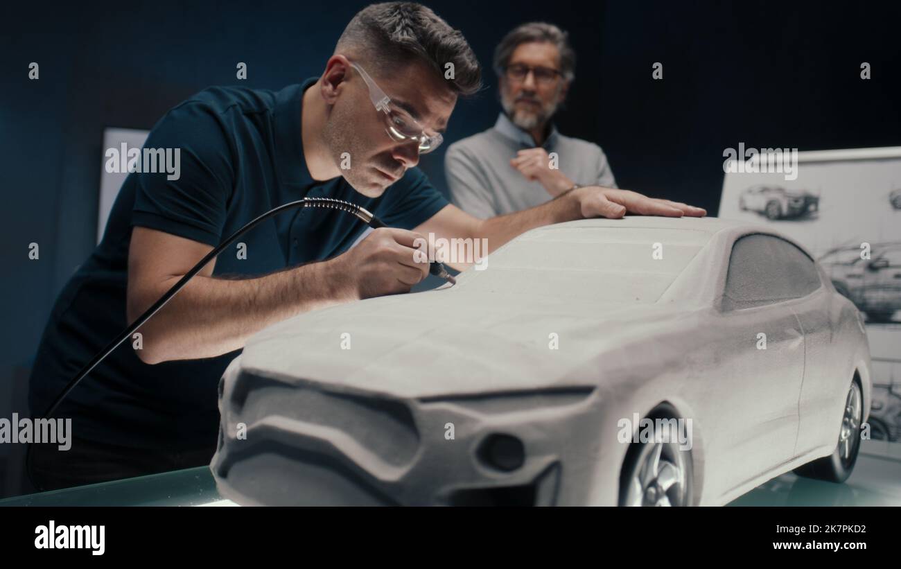 L'ingegnere automobilistico lavora sulla scultura di un prototipo di auto utilizzando uno strumento rotante mentre discute con il suo collega senior. Azienda di design di auto high tech con tecnologie innovative. Foto Stock