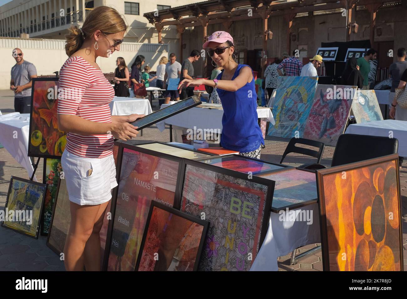 Una donna tiene opere d'arte incorniciate in una fiera d'arte all'aperto nel quartiere storico di al Fahidi. L'artista femminile dietro il tavolo indica e parla del suo lavoro. Foto Stock