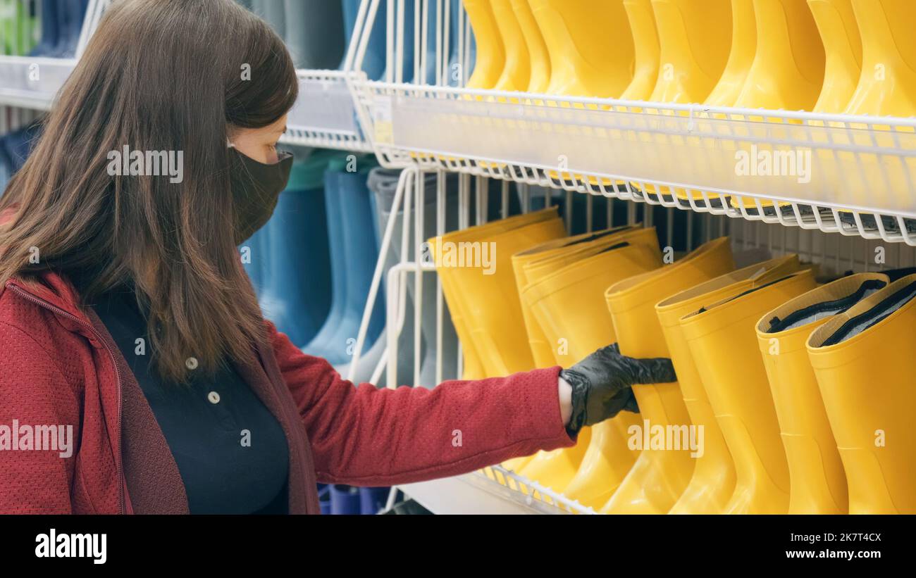 Donna caucasica che indossa maschera e guanti contro il virus acquista stivali di gomma in negozio Foto Stock