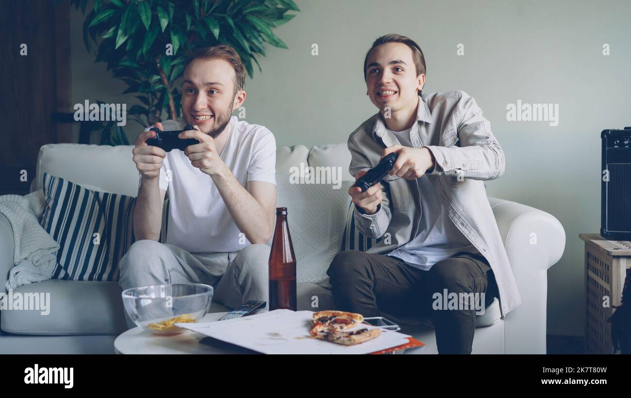 Due bei ragazzi stanno giocando a un videogioco con joystick seduti sul divano di casa. Gli uomini emotivi stanno godendo di gioco, spuntini e bottiglie di birra sono visibili. Foto Stock
