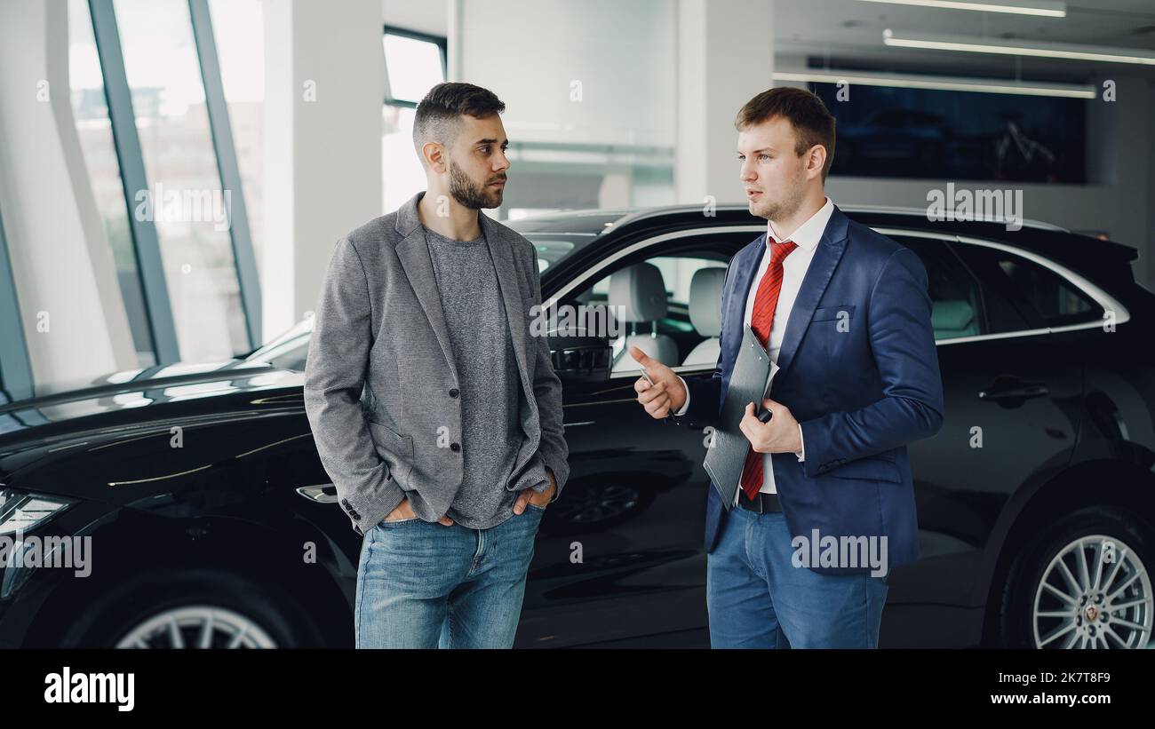 Bel ragazzo bearded sta parlando con il responsabile della concessionaria auto in mostra automobilistica discutere il nuovo modello di automobile, il concessionario è in possesso di documenti e penna. Veicolo di acquisto e concetto di persone. Foto Stock