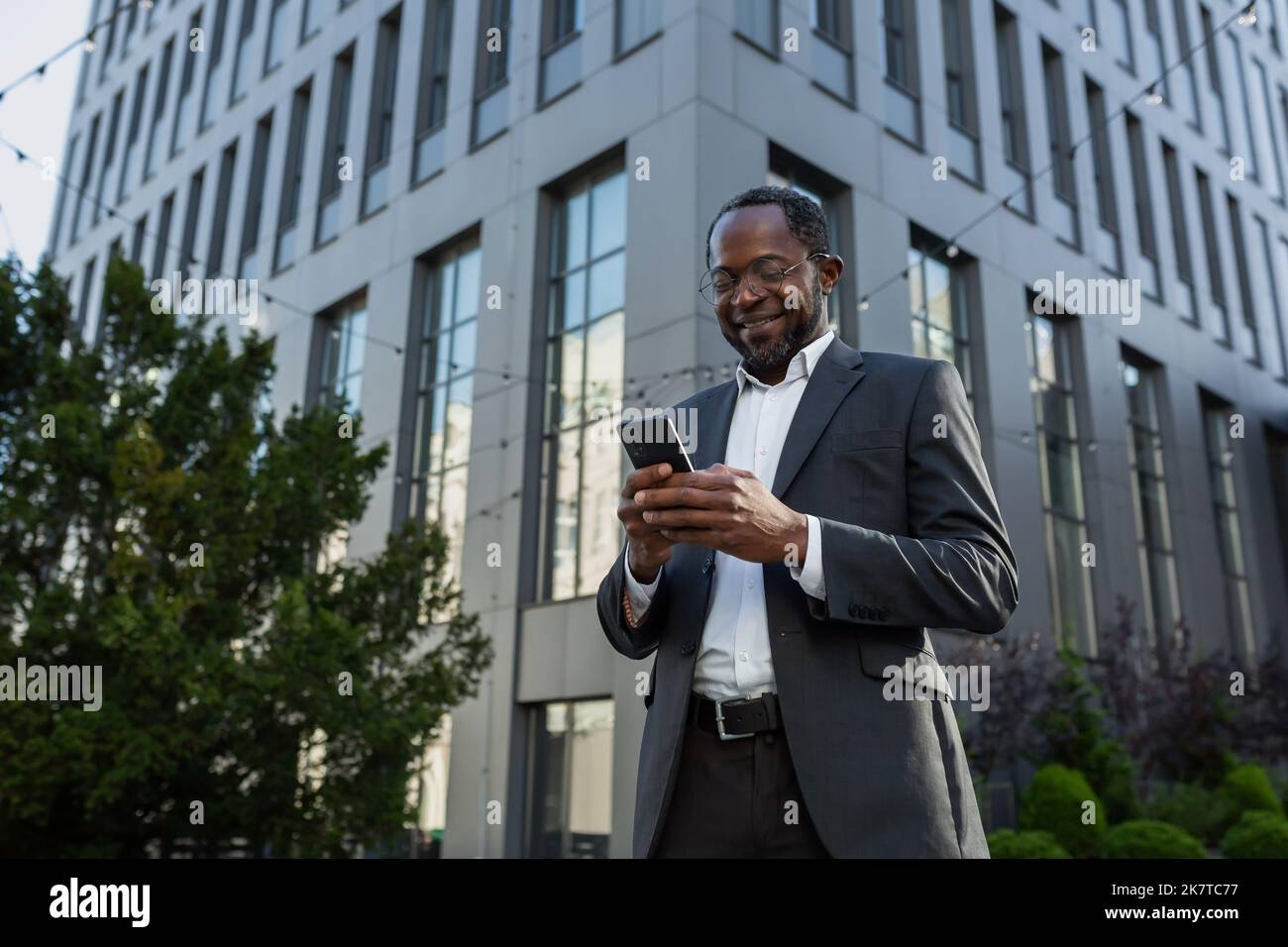 Boss afro-americano maturo usando il telefono, l'uomo d'affari fuori ufficio edificio in tuta d'affari e occhiali, uomo anziano che digita il messaggio e la navigazione pagine online sorridendo. Foto Stock