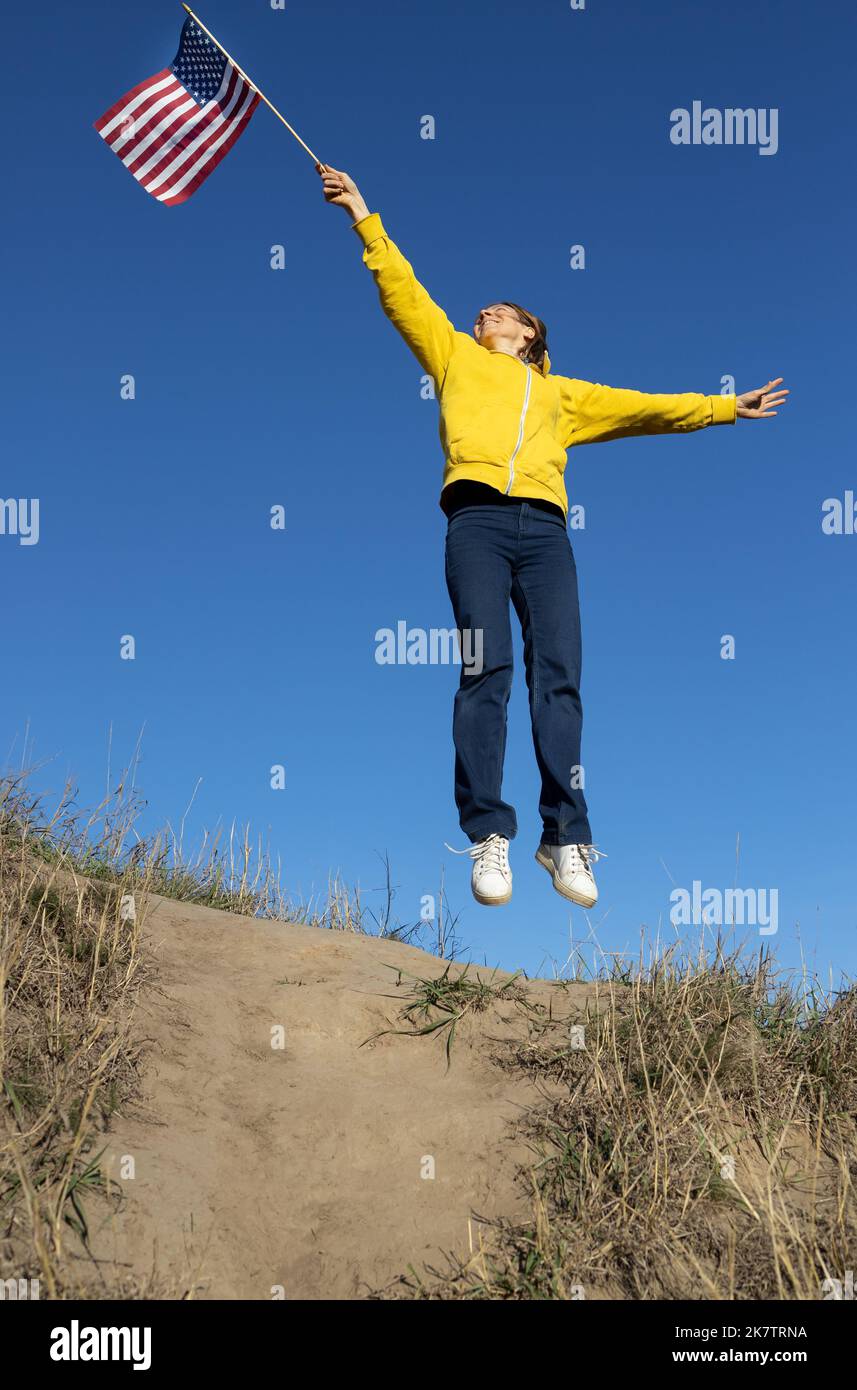 felice donna attiva jump in su alzando bandiera americana contro cielo blu nuvoloso, 4th luglio celebrazione. Independence Day degli Stati Uniti di A. Foto Stock