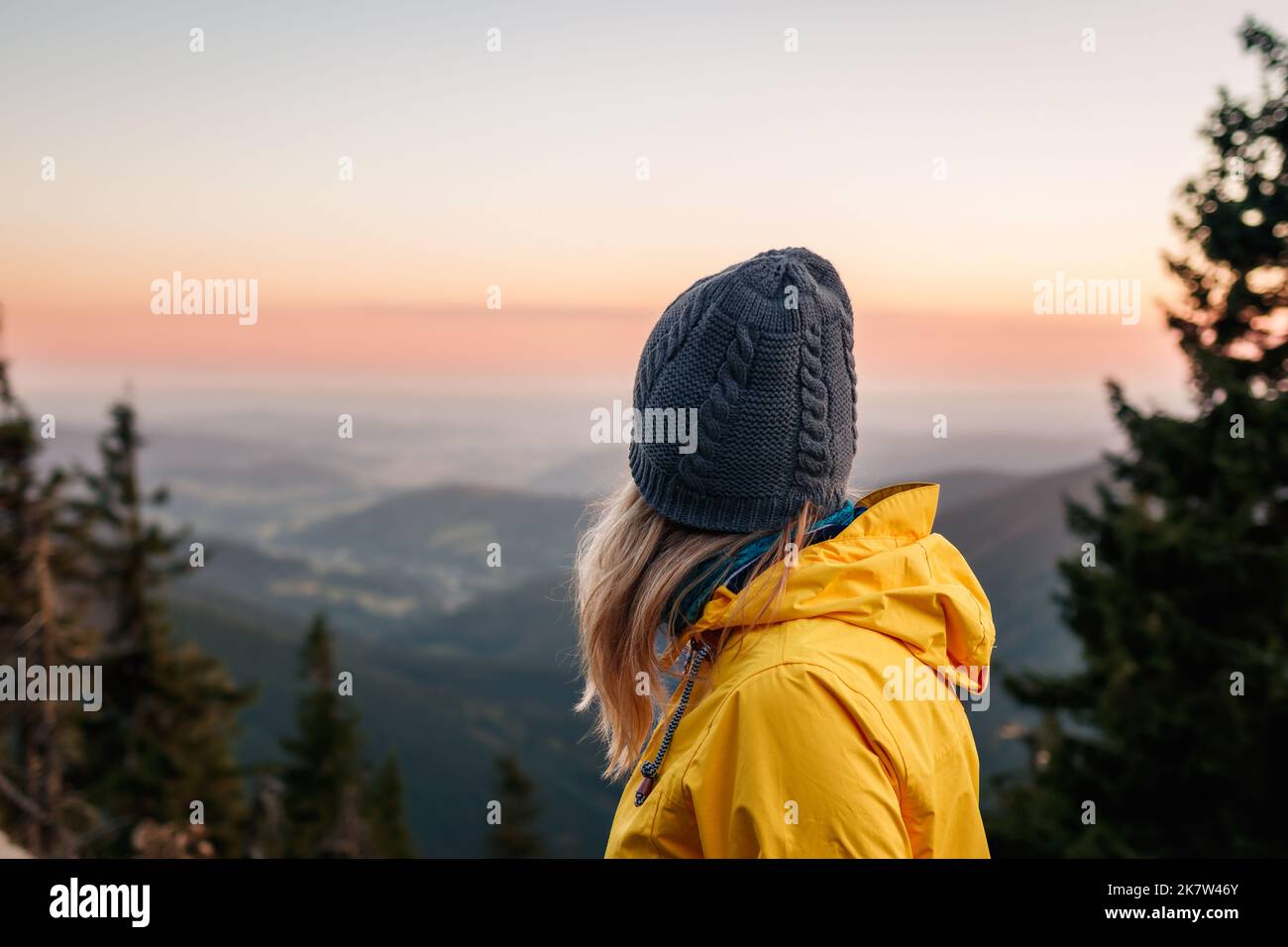 Donna con cappello a maglia e giacca gialla che guarda la catena montuosa durante il tramonto. Relax durante le escursioni in montagna Foto Stock