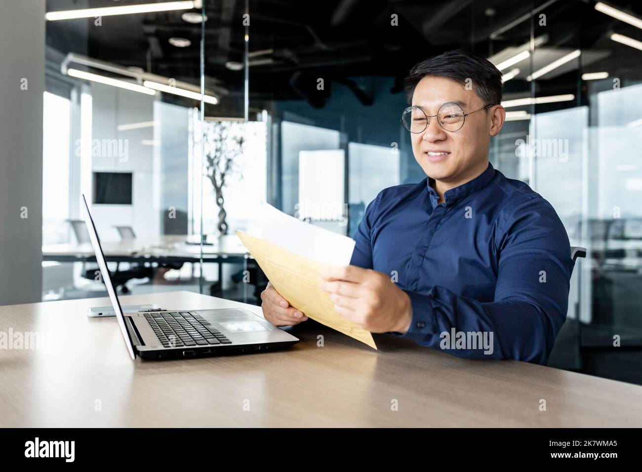 Felice uomo asiatico in occhiali ricevuti lettera apre e legge il messaggio documento, l'uomo d'affari lavora all'interno di un moderno edificio ufficio sul computer portatile, buone notizie nella notifica. Foto Stock