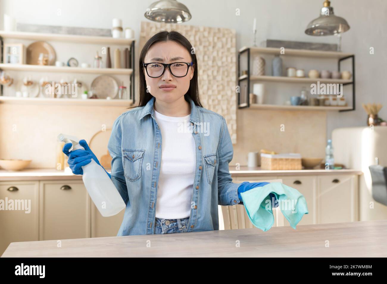 Ritratto di insoddisfatto giovane donna asiatica, casalinga in occhiali guardando la macchina fotografica tenendo detergente e spugna. Foto Stock