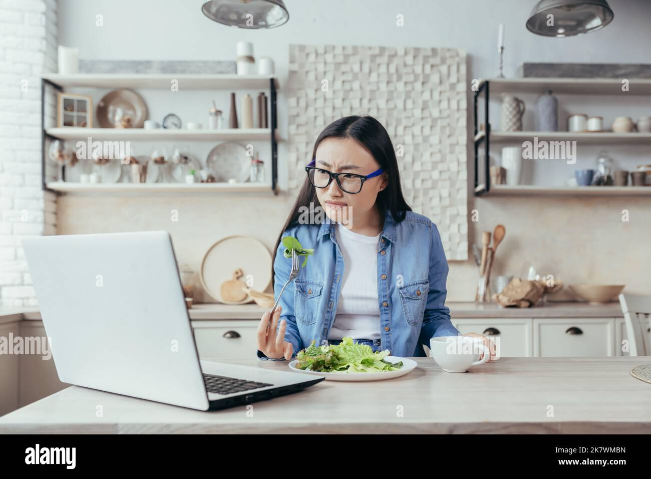 Una donna asiatica interessata è seduta al tavolo da cucina a casa. Guarda con attenzione il seminario online sulla dietetica e sul mangiare sano. Mangia un'insalata fresca, tiene una tazza con un drink in mano. Foto Stock