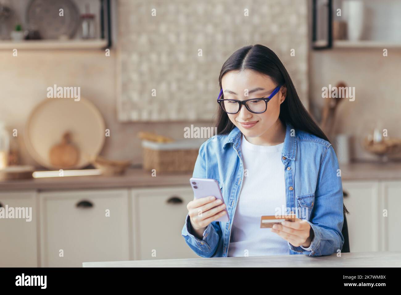 Una giovane bella donna asiatica in occhiali e una camicia denim tiene un telefono e una carta di credito in mano, usa. Si siede a casa in cucina, fa acquisti online, fa un ordine, smilling. Foto Stock