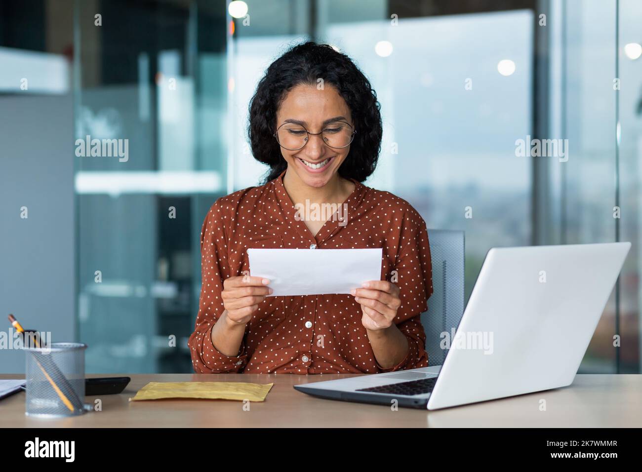 Businesswoman ha ricevuto felice lettera di notizie dalla banca, donna ispanica con capelli ricci e occhiali lavora all'interno di un moderno edificio ufficio, utilizza il computer portatile. Foto Stock