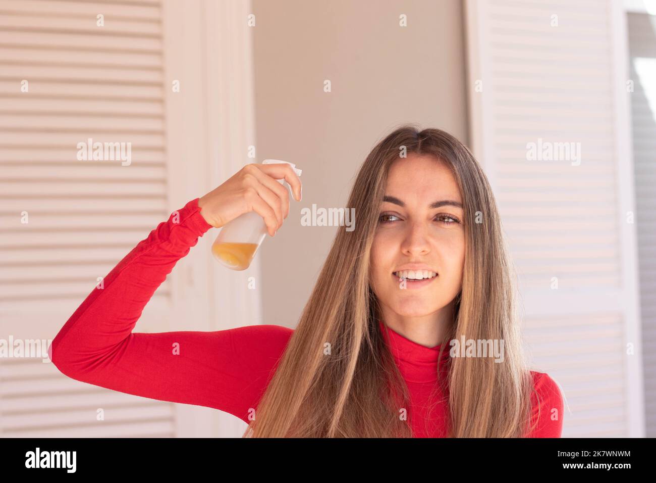 Vista frontale della giovane bella donna spruzzando oli essenziali da una bottiglia di vetro nei suoi capelli lunghi sani guardando via Foto Stock