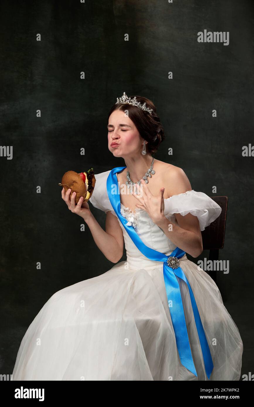Godetevi il gusto. Bella giovane regina o principessa in vestito medievale bianco mangiare hamburger su sfondo vintage scuro. Arte, creatività, bellezza, gusto Foto Stock