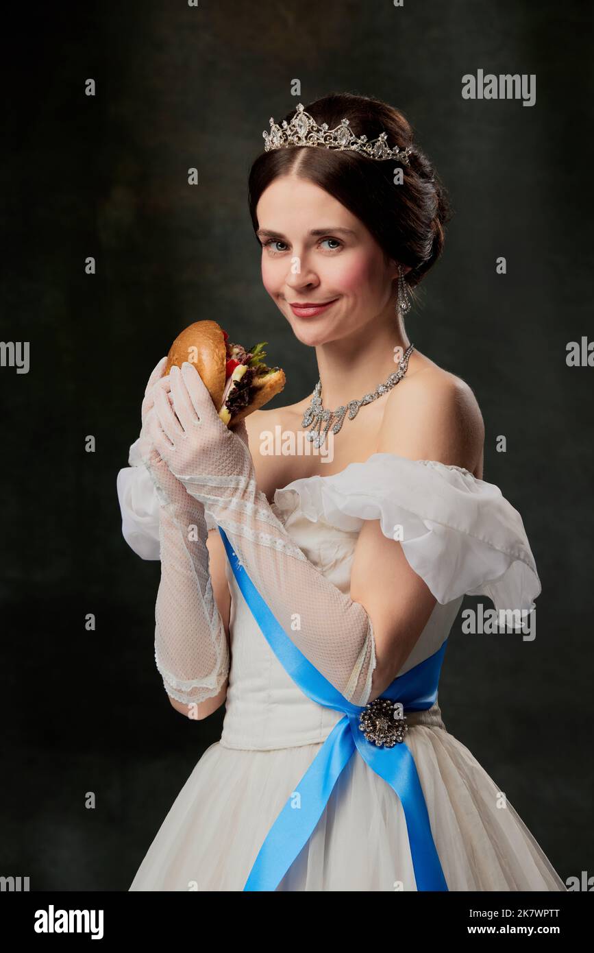 Invito al gusto. Giovane donna affascinante, persona reale, regina o principessa in vestito bianco medievale con succoso hamburger su sfondo scuro. Foto Stock