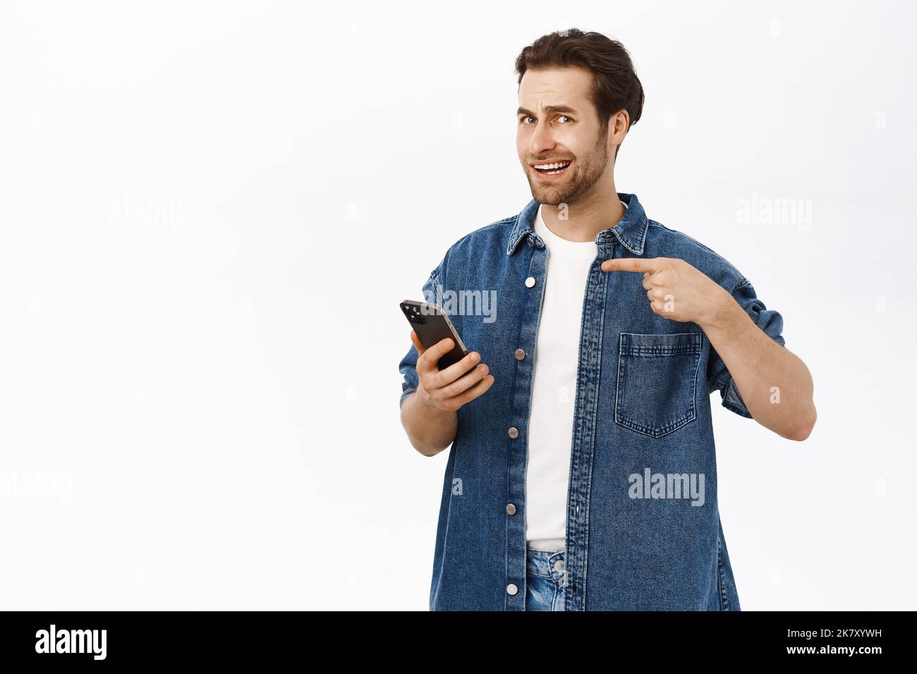 Ritratto dell'uomo con smartphone, punta al telefono cellulare con il volto scettico e deluso, in piedi su sfondo bianco Foto Stock