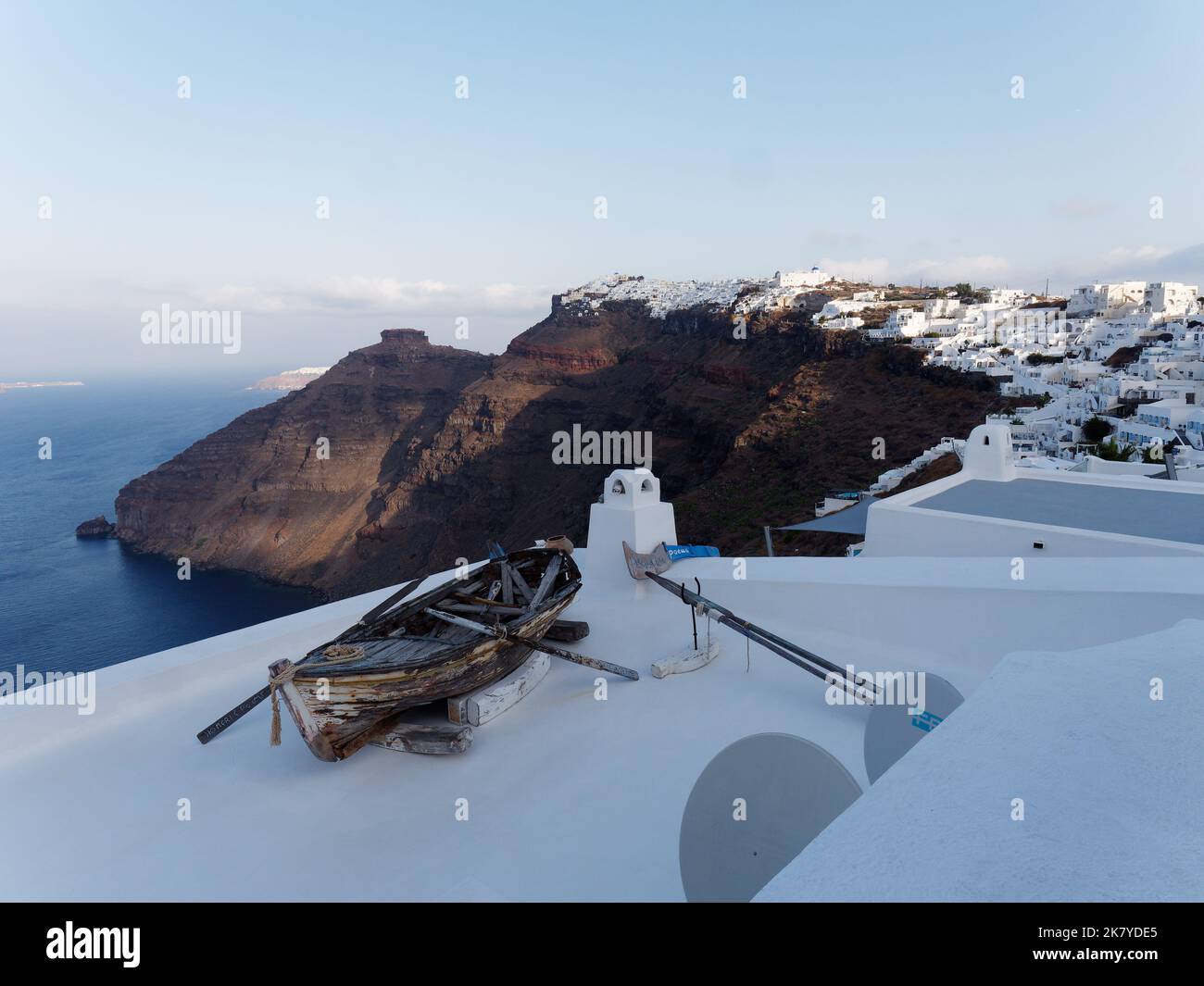 Vista verso la roccia di Skaros e la città di Imerovigli sull'isola greca delle Cicladi di Santorini nel Mar Egeo. Barca in legno sul tetto in primo piano. Foto Stock