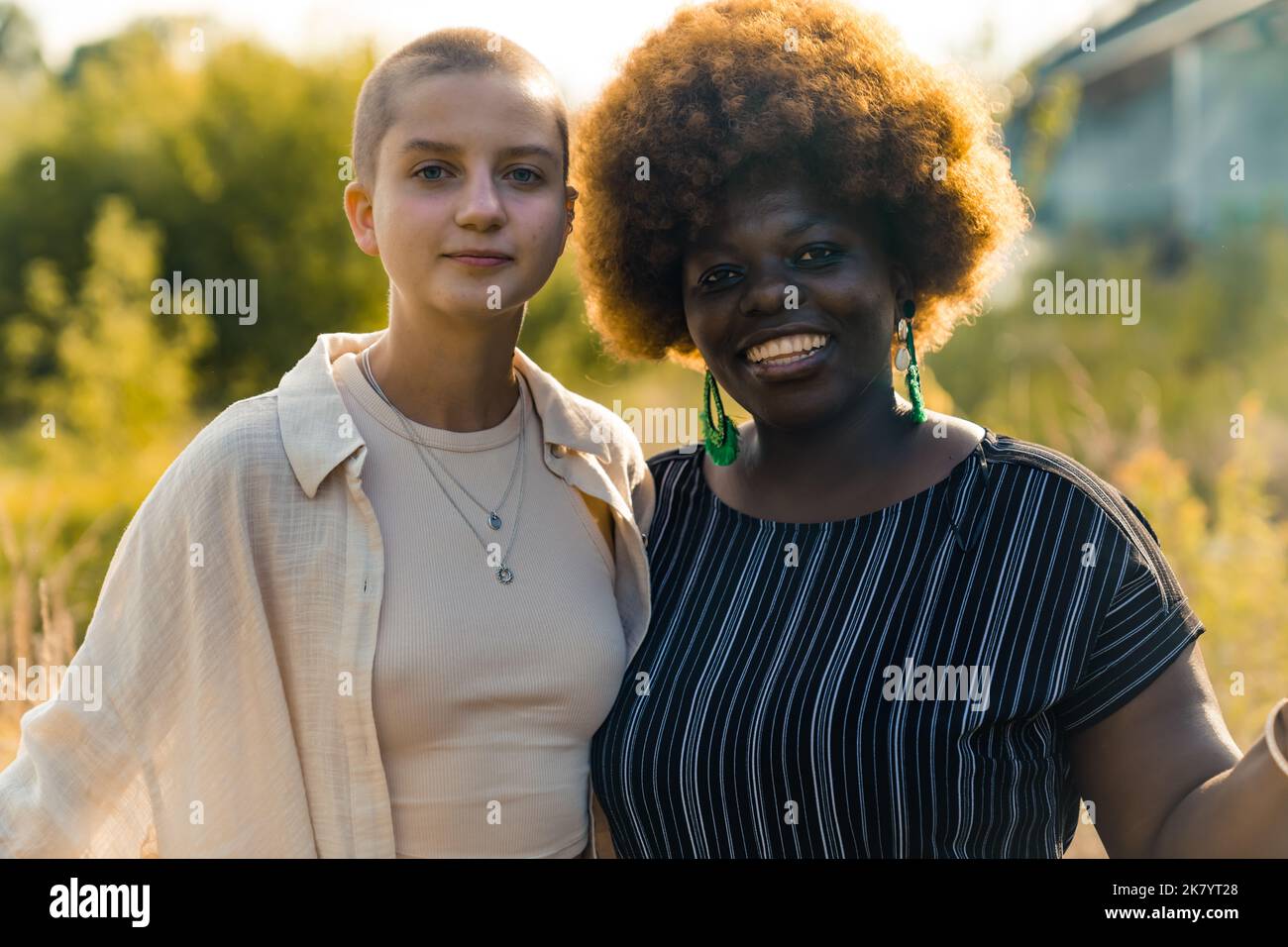 Uguaglianza nelle relazioni umane. Bel legame tra due persone queer omosessuali - donna femminile nera positiva con acconciatura afro e calva caucasica skinny non-binaria persona. Ripresa all'aperto con sfondo sfocato. Sole. Foto di alta qualità Foto Stock