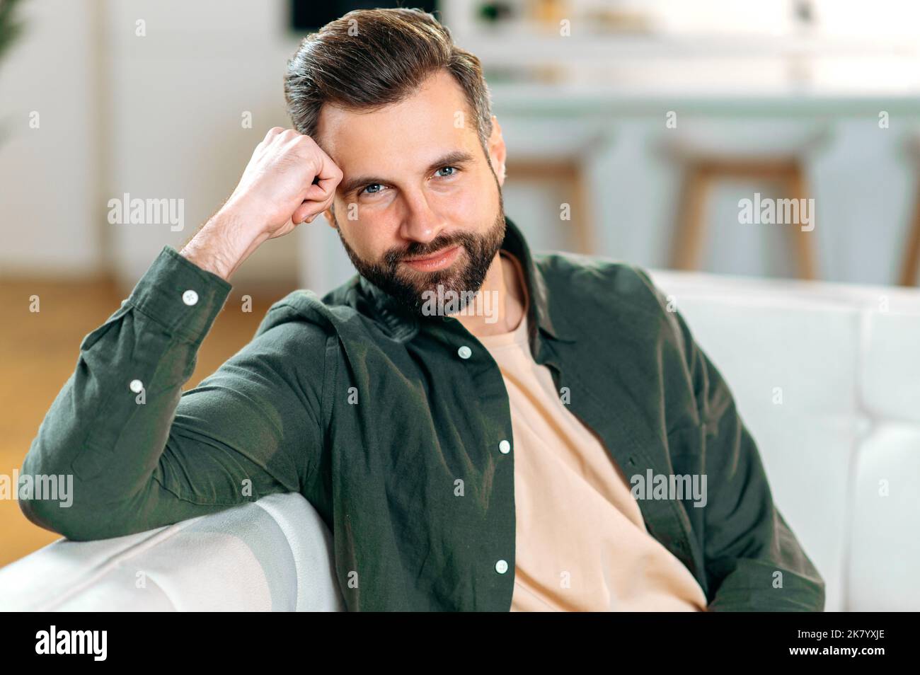 Ritratto maschile. Foto di un attraente uomo caucasico moderno e positivo, in abiti casual elegante, si siede su un divano nel soggiorno, guarda la macchina fotografica, sorride amichevole Foto Stock