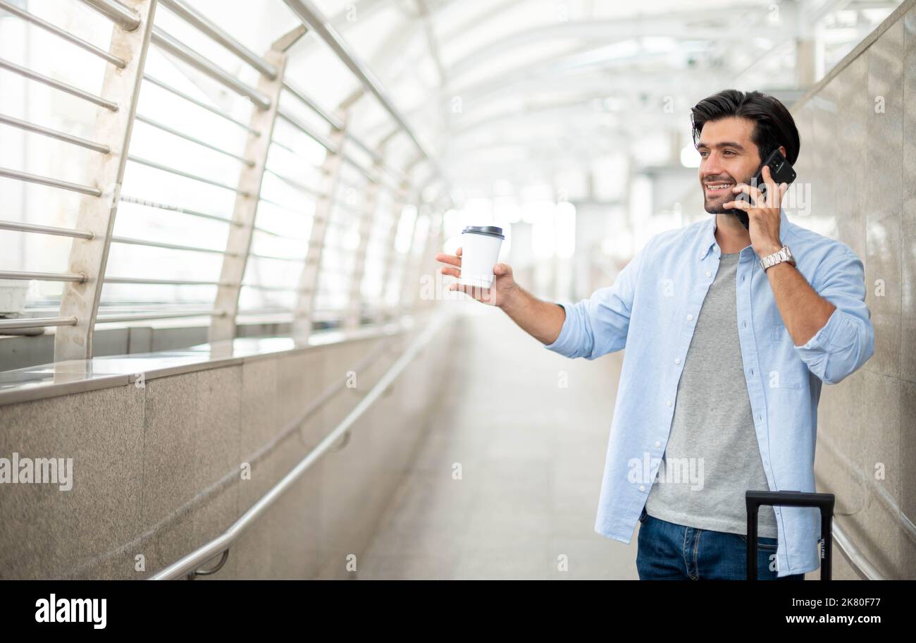 L'uomo utilizza lo smartphone mentre l'altra mano tiene una tazza di caffè e tira i bagagli in aeroporto. Foto Stock