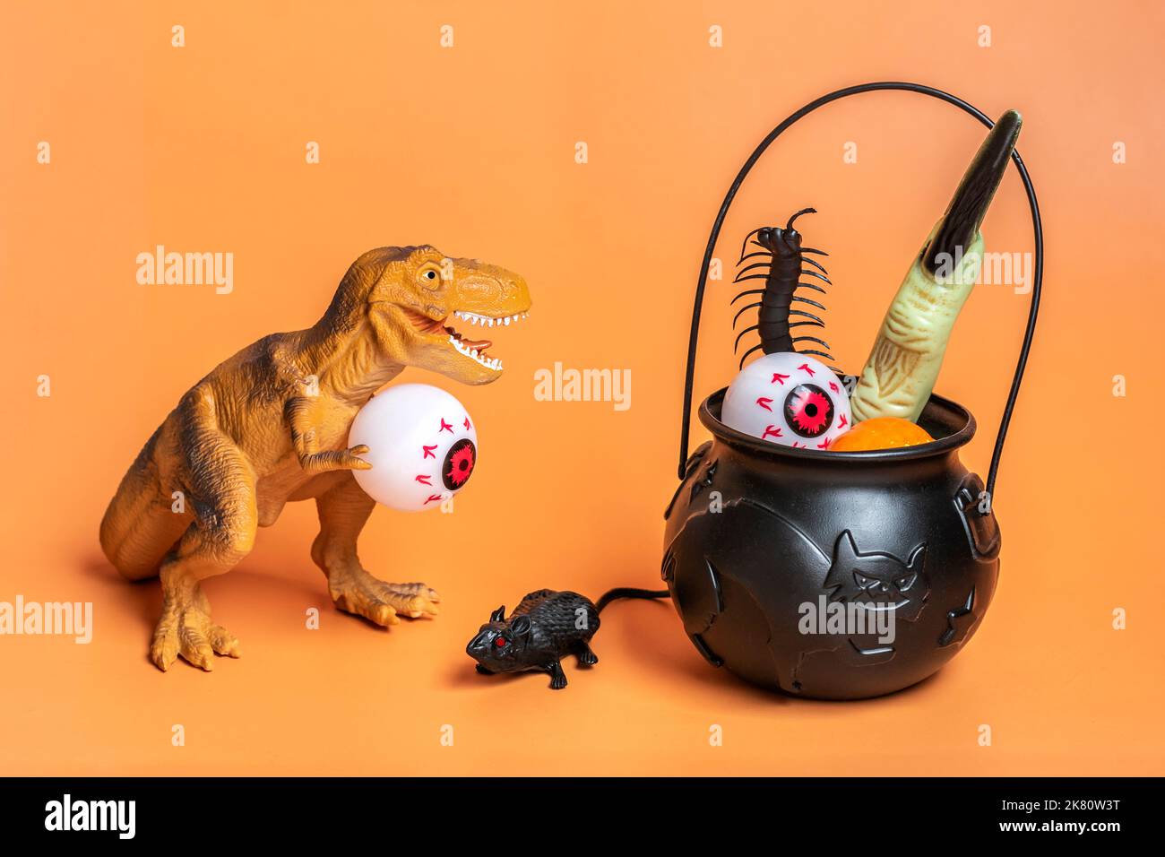Giocattolo dinosauro Tyrannosaurus tenere occhio nelle sue zampe, ciotola con pipistrello, zucca isolato su sfondo arancione festivo auguri carta di Halloween felice giorno cre Foto Stock