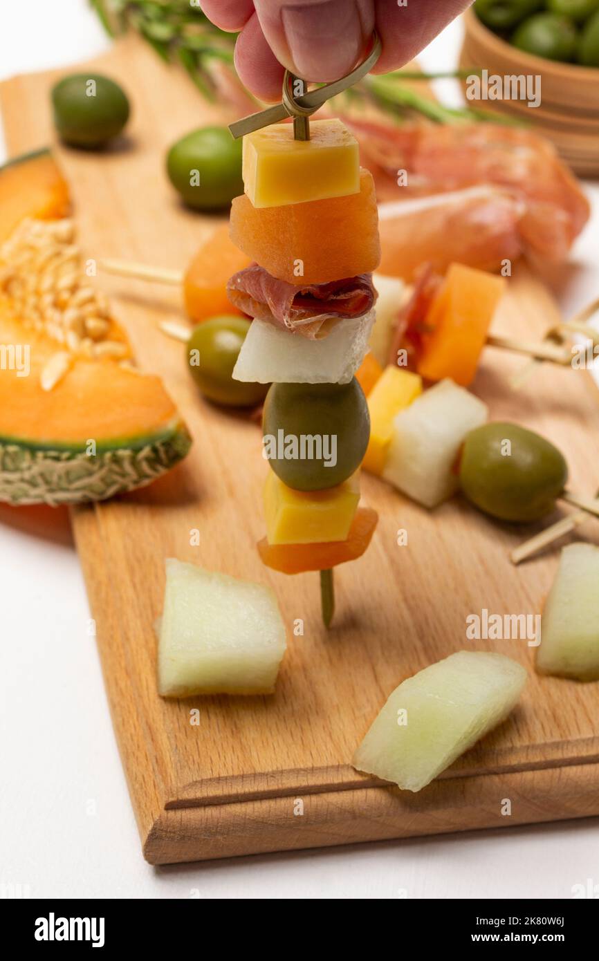 Si tiene a mano spiedino con formaggio, olive e prosciutto. Melone, jamon e olive a bordo. Primo piano. Sfondo bianco. Foto Stock