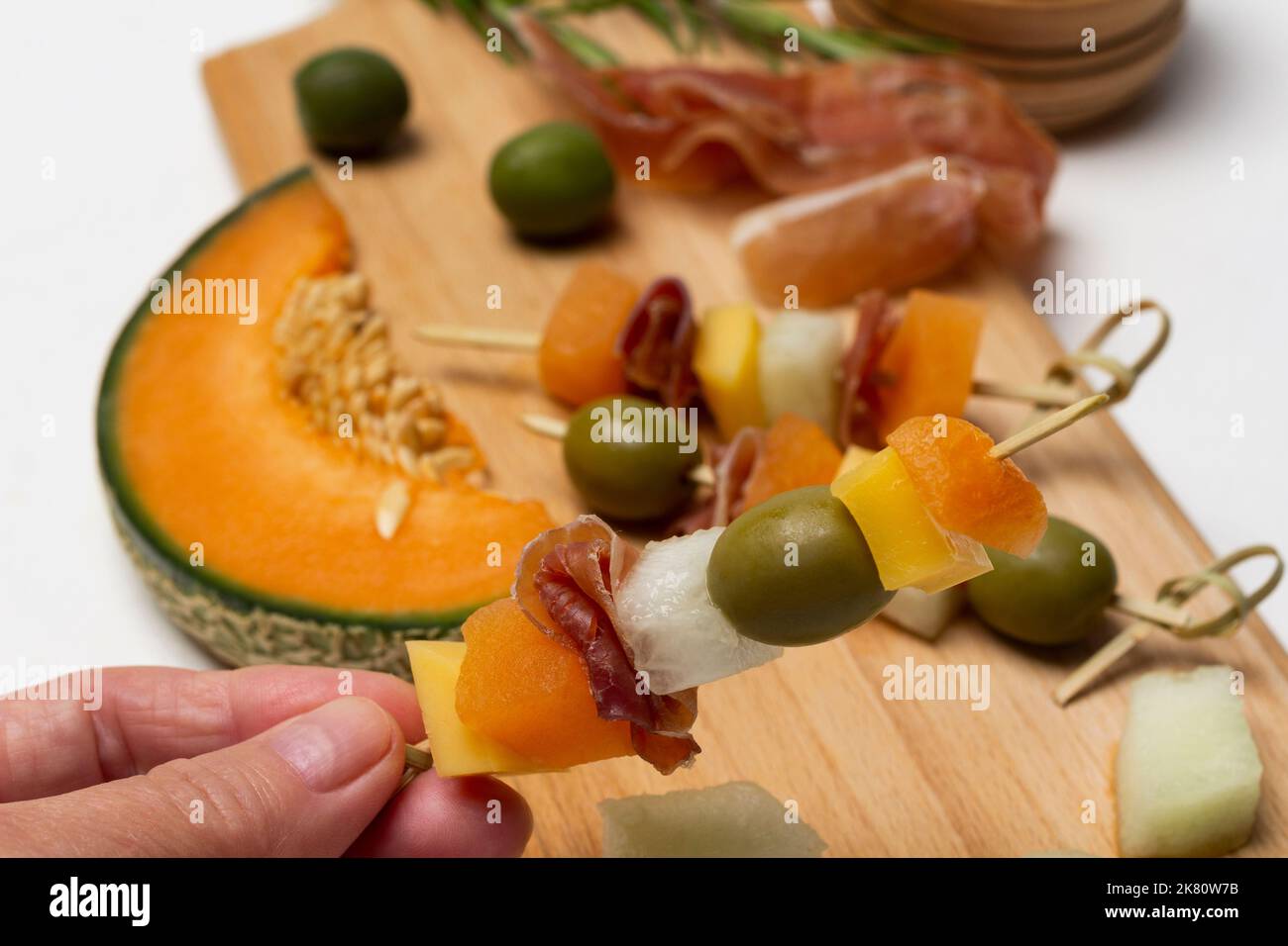 La mano tiene lo spiedino con formaggio, olive e prosciutto. Melone, jamon e olive a bordo. Primo piano. Sfondo bianco. Foto Stock