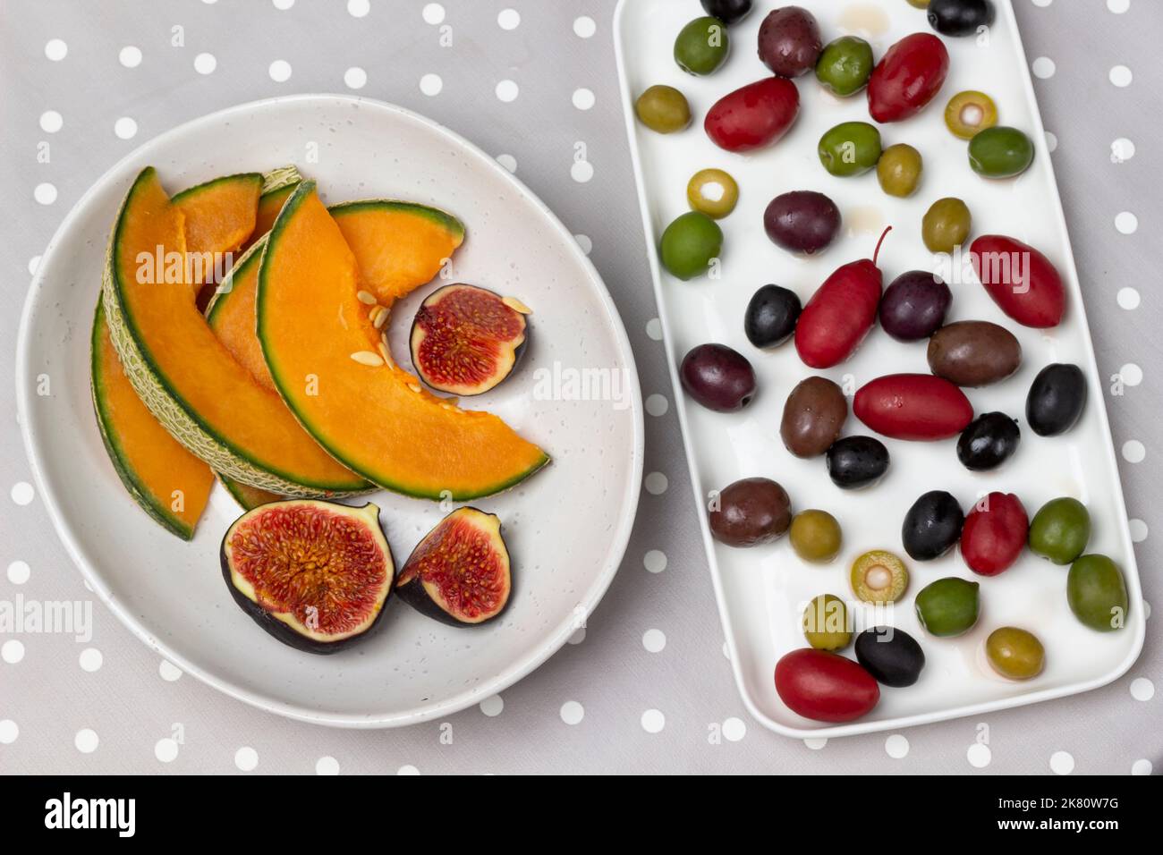 Olive di varie varietà su piatto, fettine di melone e fichi tagliati in ciotola. Giacitura piatta. Sfondo a pois grigio. Foto Stock