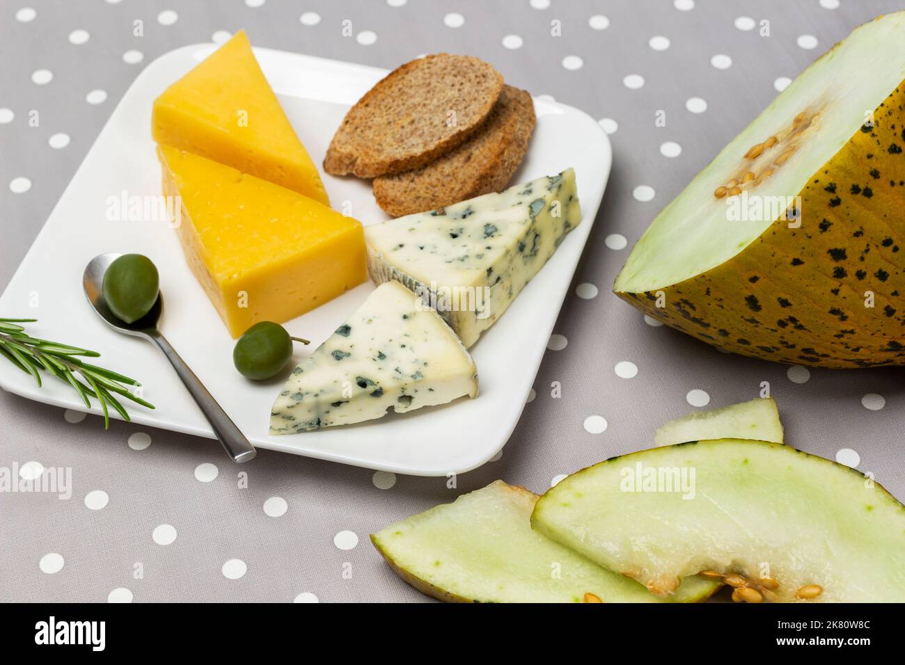 Melone, diversi tipi di formaggio e olive su piatto. Vista dall'alto. Sfondo a pois grigio. Foto Stock