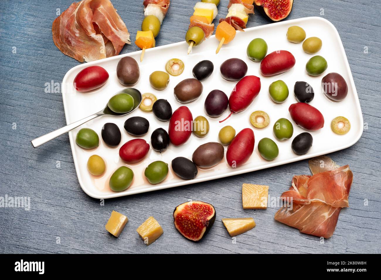 Jamon, formaggio, fichi e spiedini con formaggio, olive e jamon sul tavolo. Olive di varie varietà su piatto. Giacitura piatta. Sfondo blu. Foto Stock