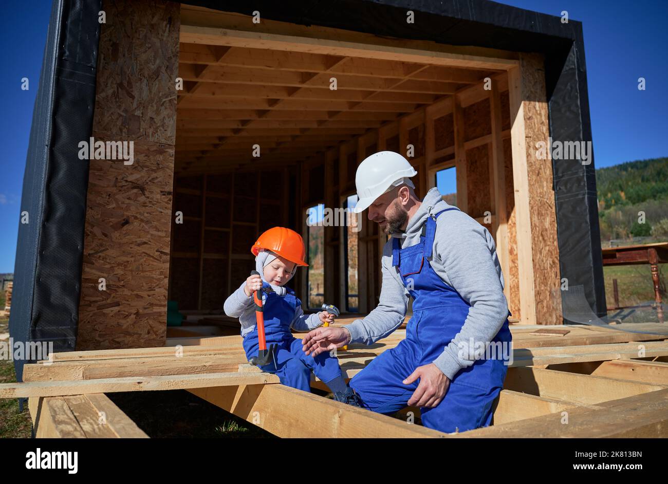 Padre con bambino figlio costruzione legno cornice casa. Ragazzo che aiuta il suo papà, giocando con il bambino in cantiere, indossando casco e tute blu nella giornata di sole. Carpenteria e concetto di famiglia. Foto Stock