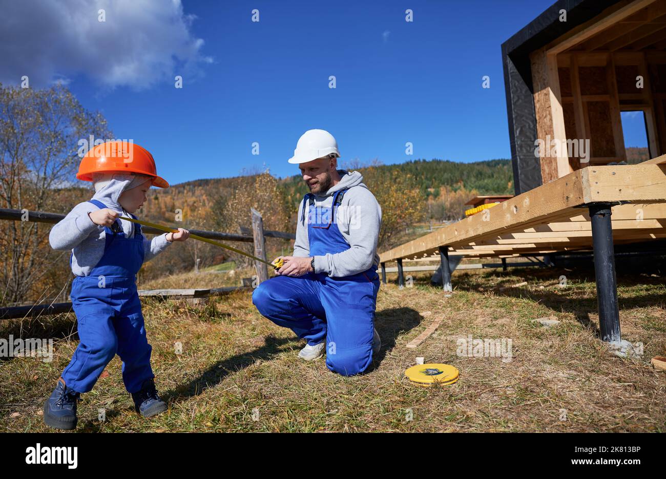 Padre con figlio piccolo costruendo casa con struttura in legno. Ragazzo che aiuta il suo papà, giocando con metro in cantiere, indossando casco e tute blu nelle giornate di sole. Falegnameria e concetto di famiglia. Foto Stock