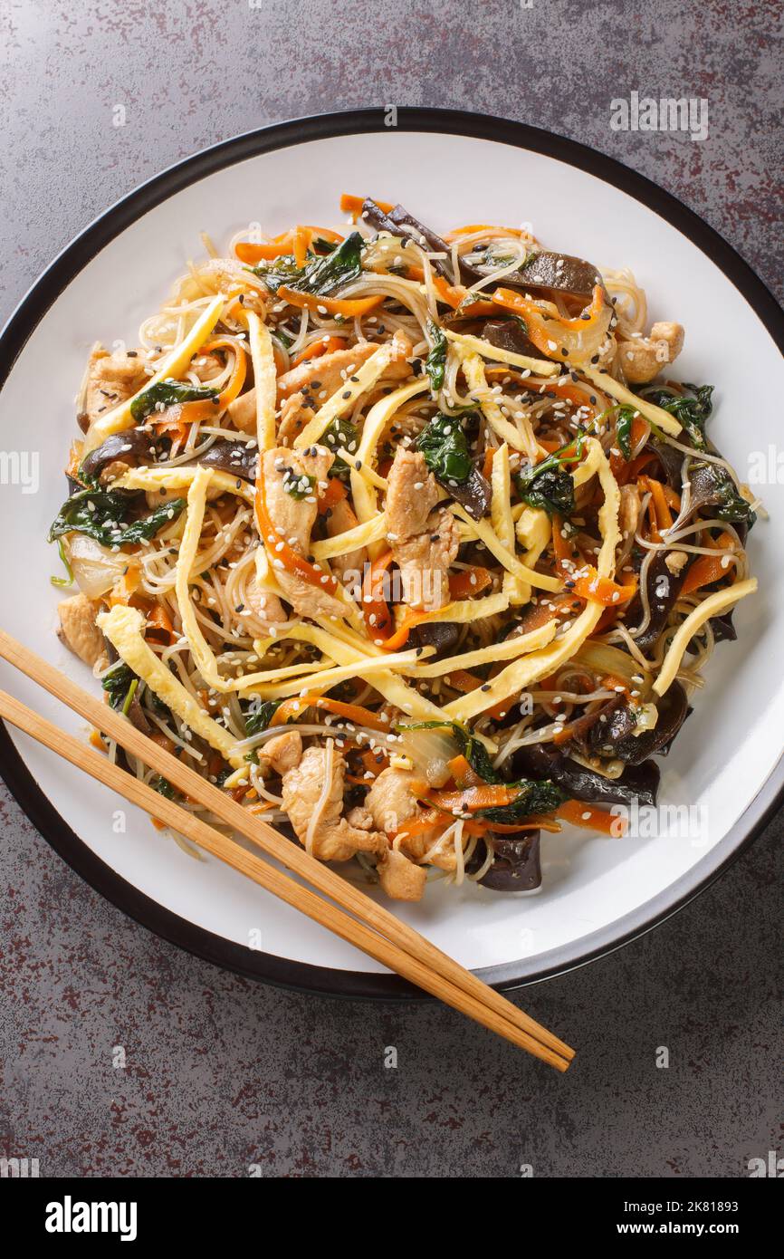 japchae o spaghetti coreani fritti con verdure e maiale conditi con il primo piano di sesamo sul piatto sul tavolo. Vista verticale dall'alto da Foto Stock
