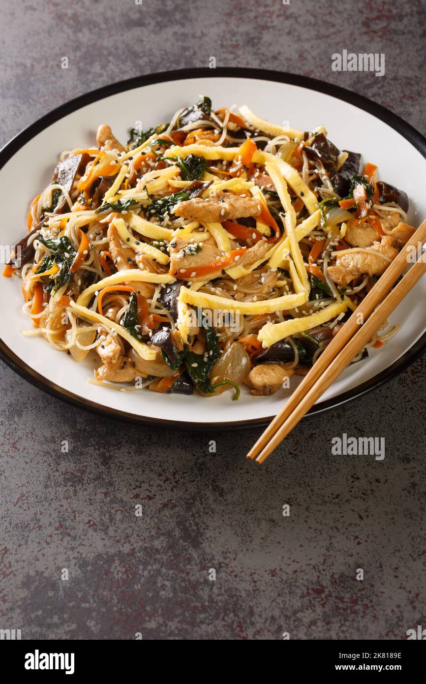 japchae o spaghetti coreani fritti con verdure e maiale conditi con il primo piano di sesamo sul piatto sul tavolo. Verticale Foto Stock
