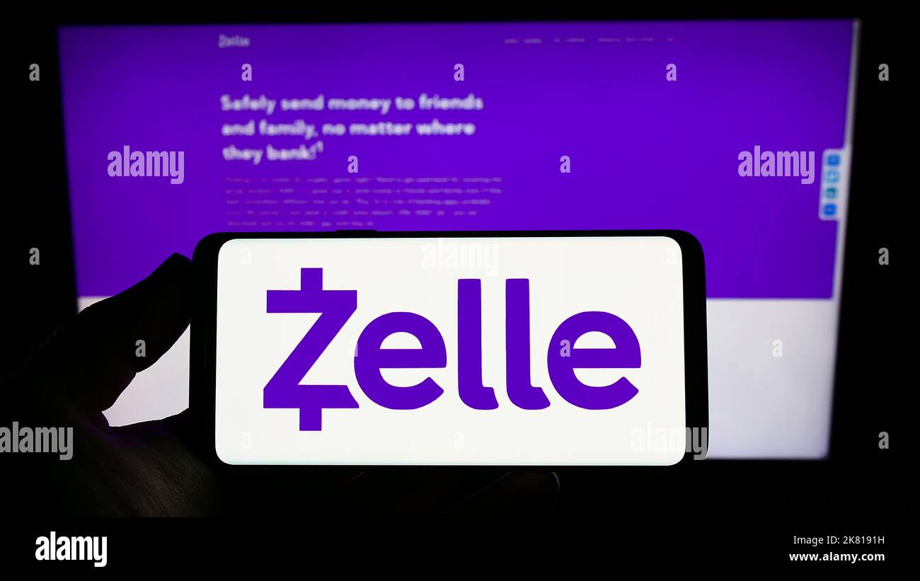 Persona che tiene in mano uno smartphone con il logo della società di pagamenti digitali USA Zelle sullo schermo di fronte al sito Web. Messa a fuoco sul display del telefono. Foto Stock