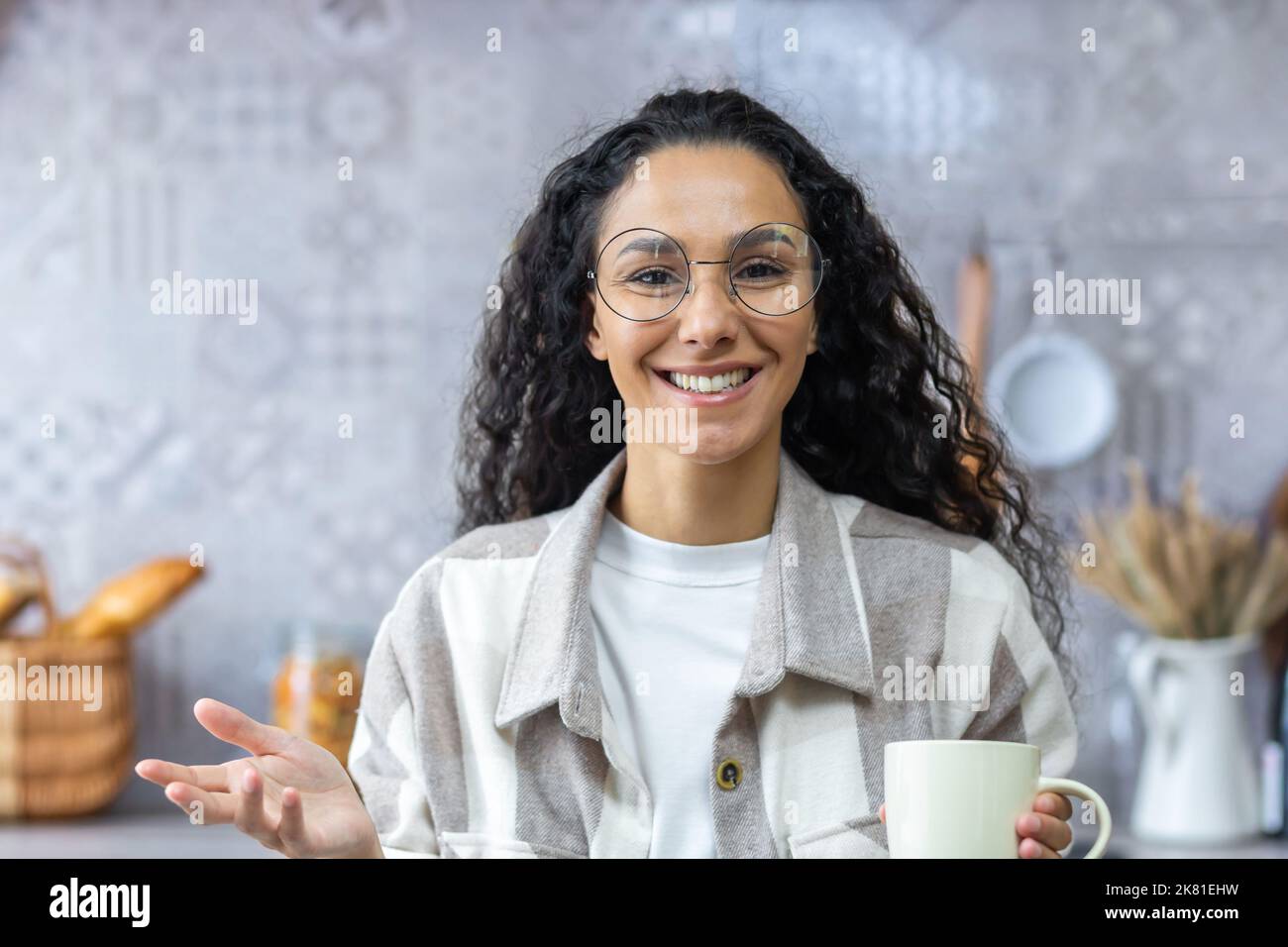 Videochiamata, comunicazione online con gli amici, felice e sorridente donna ispanica guardando la webcam e parlando, donna a casa in cucina con capelli ricci e bicchieri che tiene una tazza di bevanda calda. Foto Stock