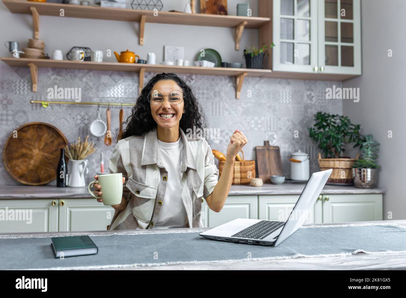 Ritratto di donna di successo a casa, donna ispanica guardando la macchina fotografica e felice celebrando trionfo vittoria, donna d'affari che utilizza il portatile per il lavoro remoto, freelance con capelli ricci e occhiali. Foto Stock