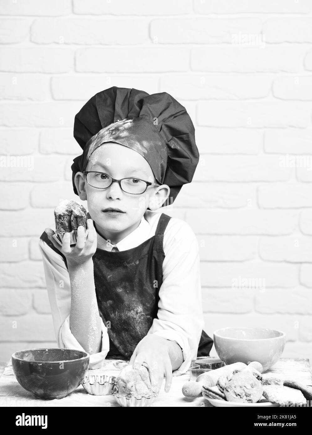 giovane ragazzo cuoco chef carino in uniforme rossa e cappello sul volto macchiato con bicchieri seduti sul tavolo con ciotole colorate, gustosi biscotti, pin rotolante e. Foto Stock