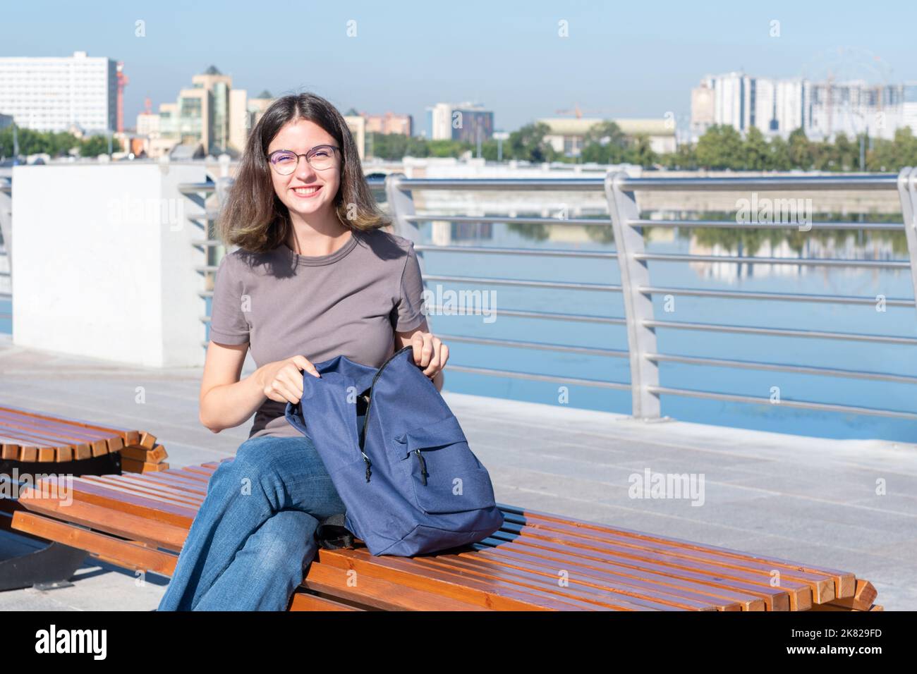 Una studentessa sorridente con gli occhiali, aprendo lo zaino, cercando qualcosa nella sua borsa, seduti all'aperto su una panca su una strada cittadina. Un adolescente s Foto Stock