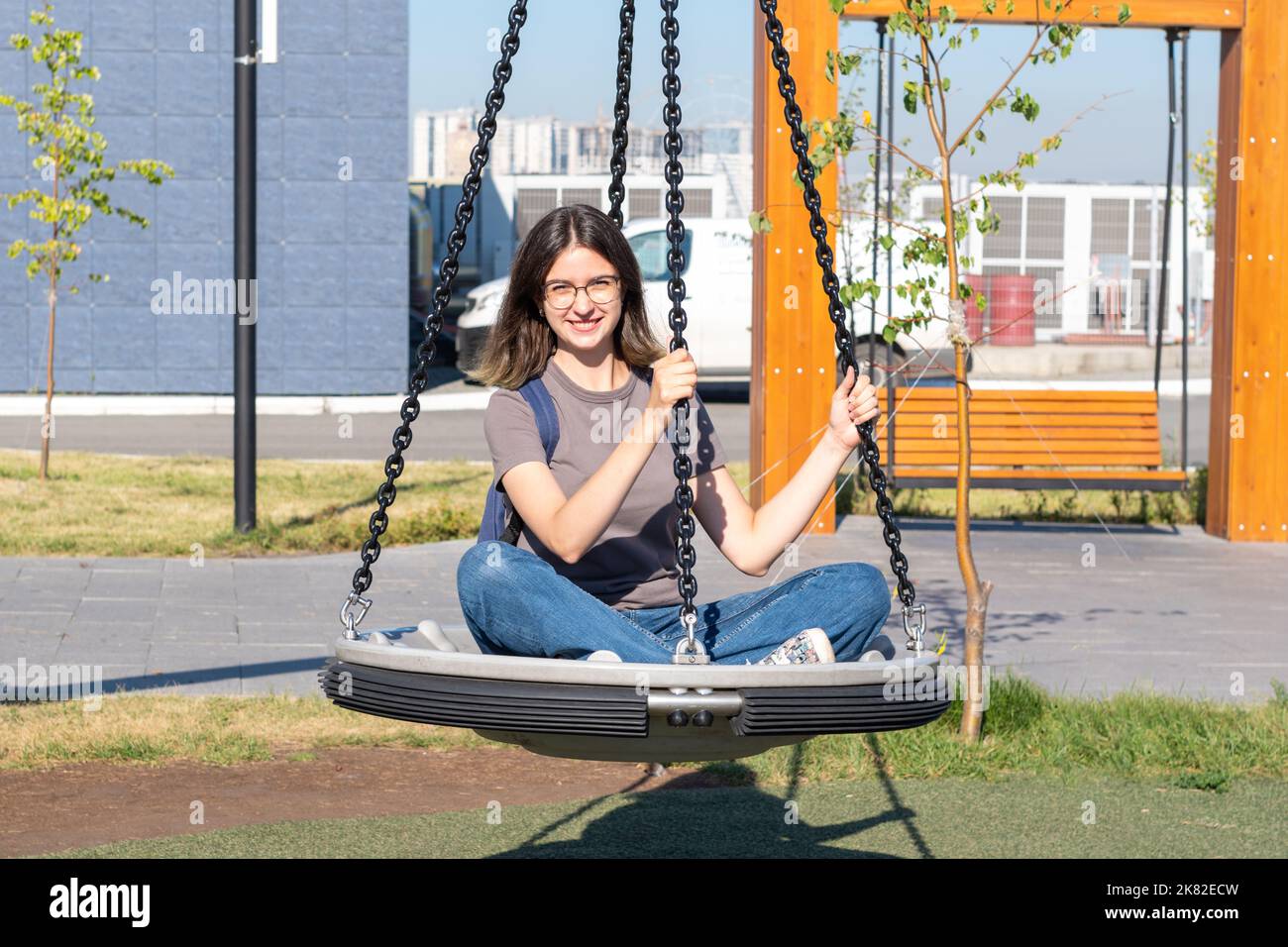 Ragazza spagnola sorridente studente in occhiali jeans che oscillano su un altalena nel parco. La ragazza si riposa dopo aver studiato. Intrattenimento estivo in città pa Foto Stock