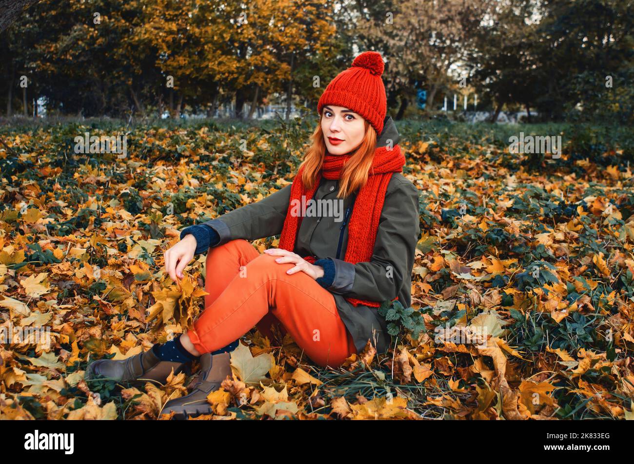 Ritratto autunnale di ragazza carina in caldo cappello a maglia arancione e sciarpa arancione seduta in foglia gialla caduta nel parco. Look romantico di grandi occhi. Guarda in Foto Stock