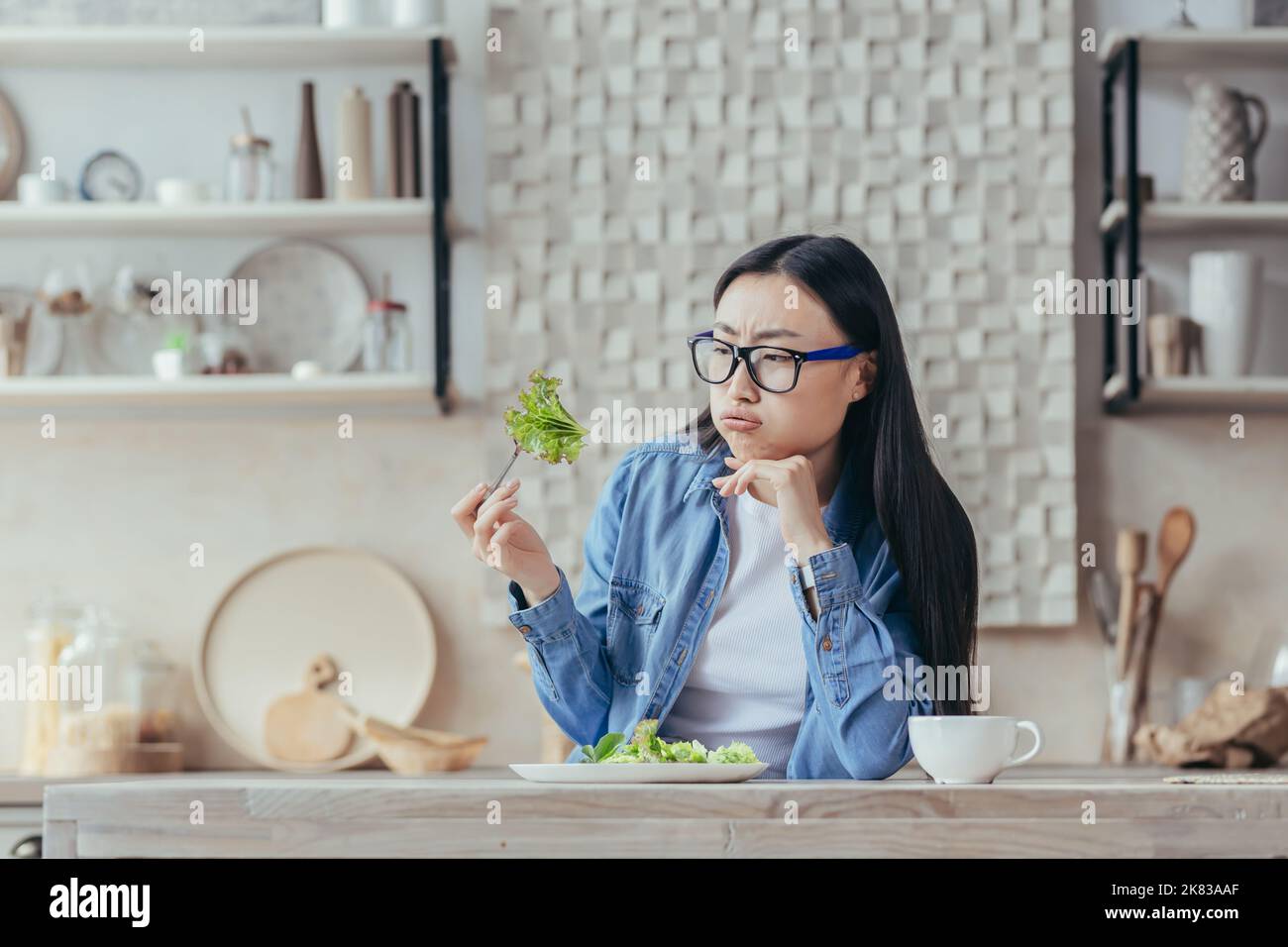 Ritratto di una giovane bella donna asiatica seduta in cucina al tavolo, tenendo una fresca insalata verde nella forchetta, sospirando, guardandola infamia. Stanco di stare, odia il vegetarismo. Foto Stock