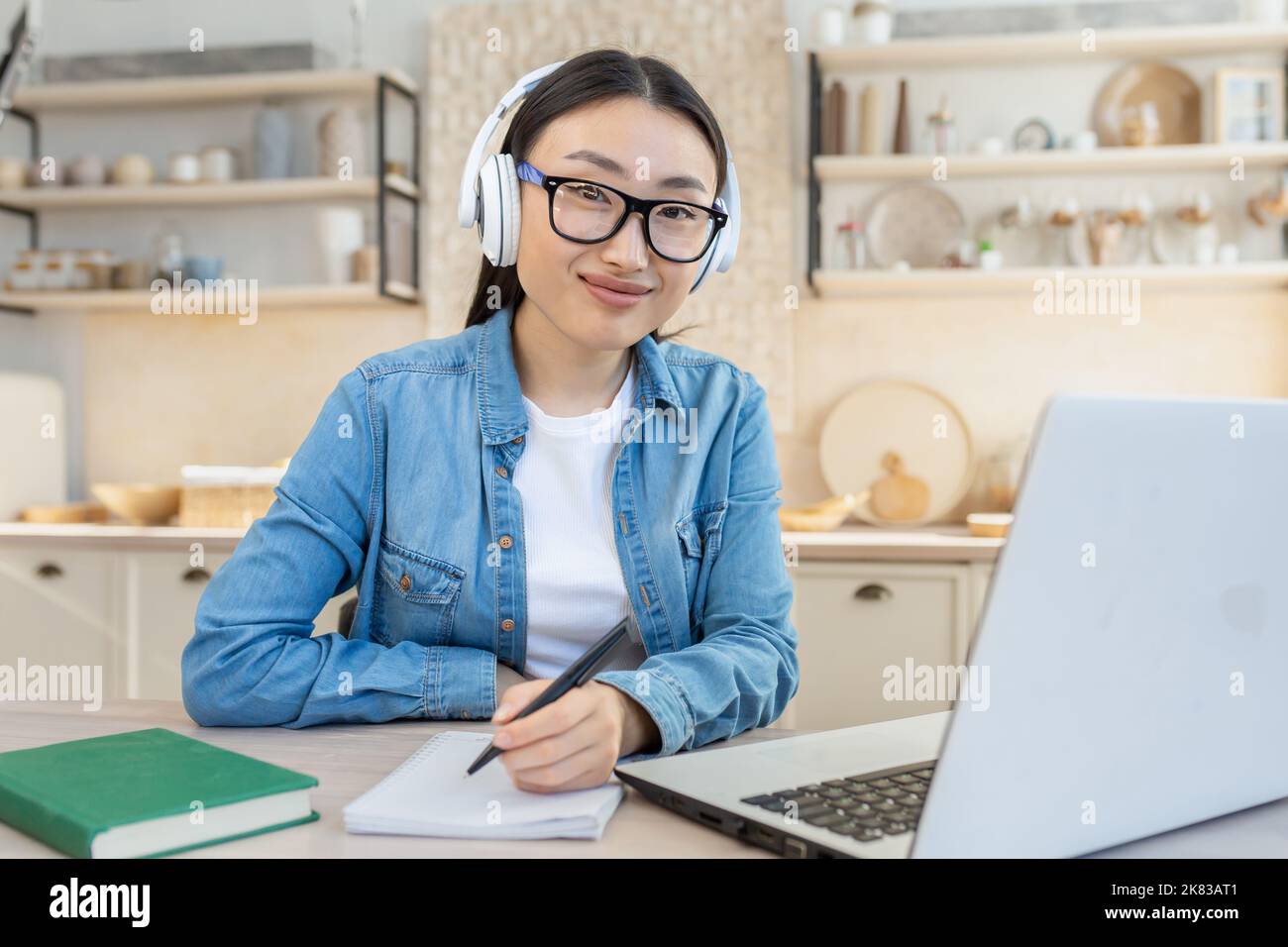 Giovane bella donna asiatica che lavora come freelancer. Seduti a casa con un computer portatile e indossando cuffie bianche, scrivendo in un notebook, guardando la fotocamera, sorridendo. Foto Stock