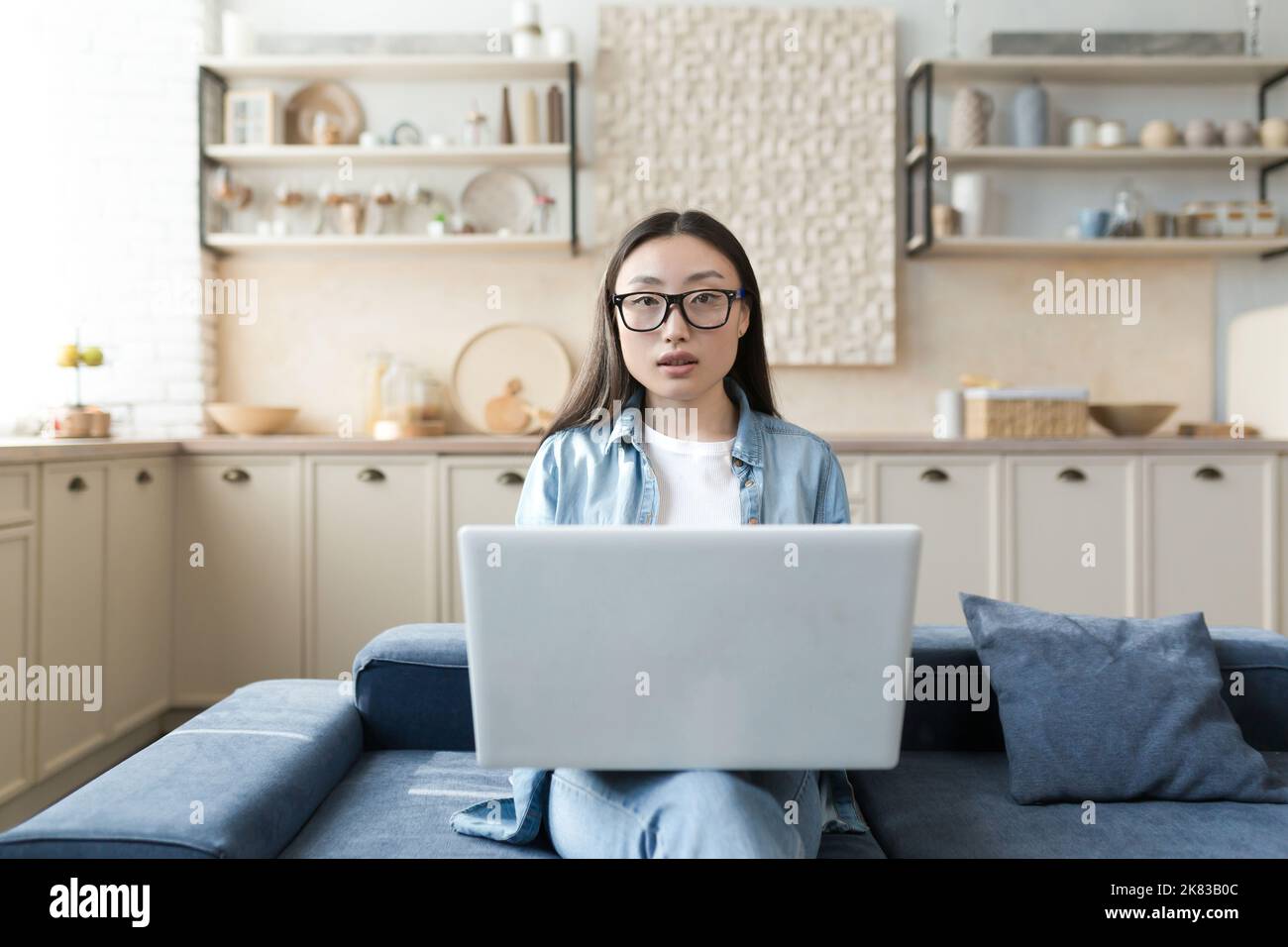 Ritratto di una giovane bella blogger femminile asiatica. Seduti a casa sul divano, tenendo un laptop bianco in grembo, guardando con intelligenza la telecamera. Foto Stock