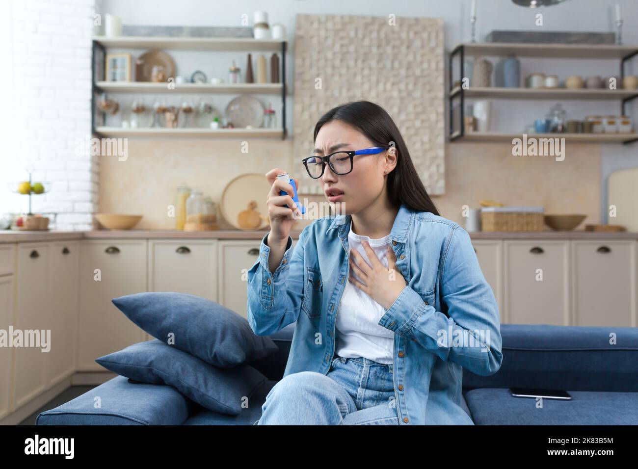 Attacco allergico. Asma. Giovane donna asiatica seduta sul divano a casa e con inalatore. Ha un attacco di asma, tratta le allergie, ha difficoltà a respirare. Foto Stock