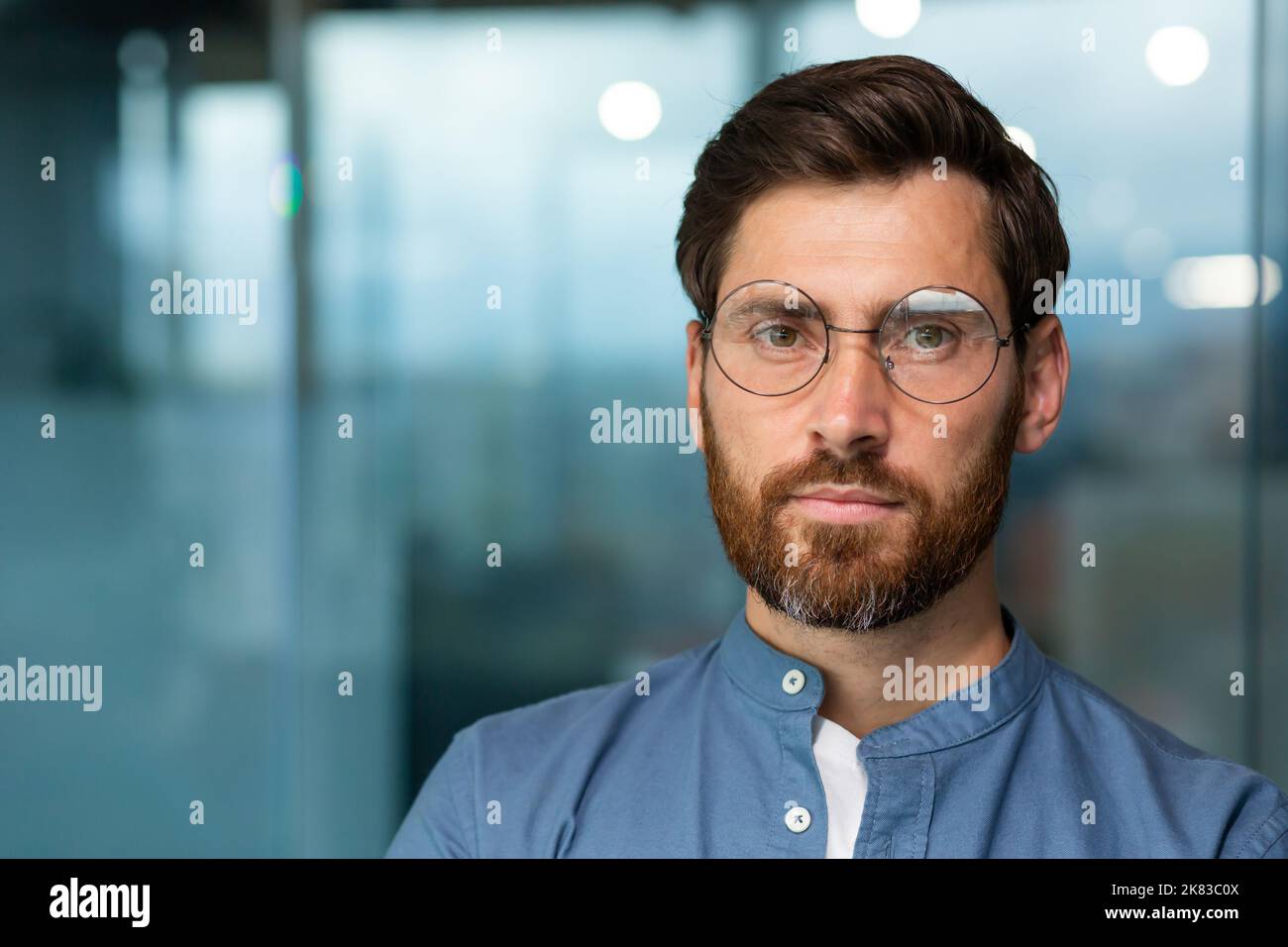 Primo piano ritratto di serio e premuroso uomo d'affari maturo con barba, uomo in camicia e occhiali guardando la macchina fotografica, startuper lavorare all'interno di un moderno edificio per uffici. Foto Stock