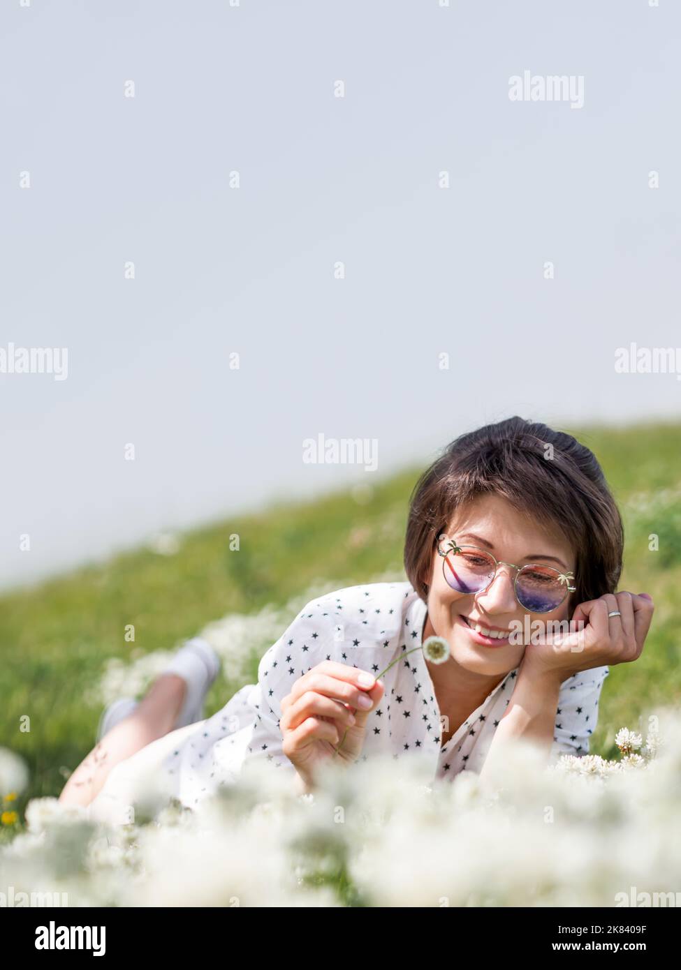 Donna in occhiali da sole colorati sniffs fiori trifoglio sul prato in parco urbano. Natura in città. Rilassati all'aperto dopo il lavoro. Atmosfera estiva. Foto Stock