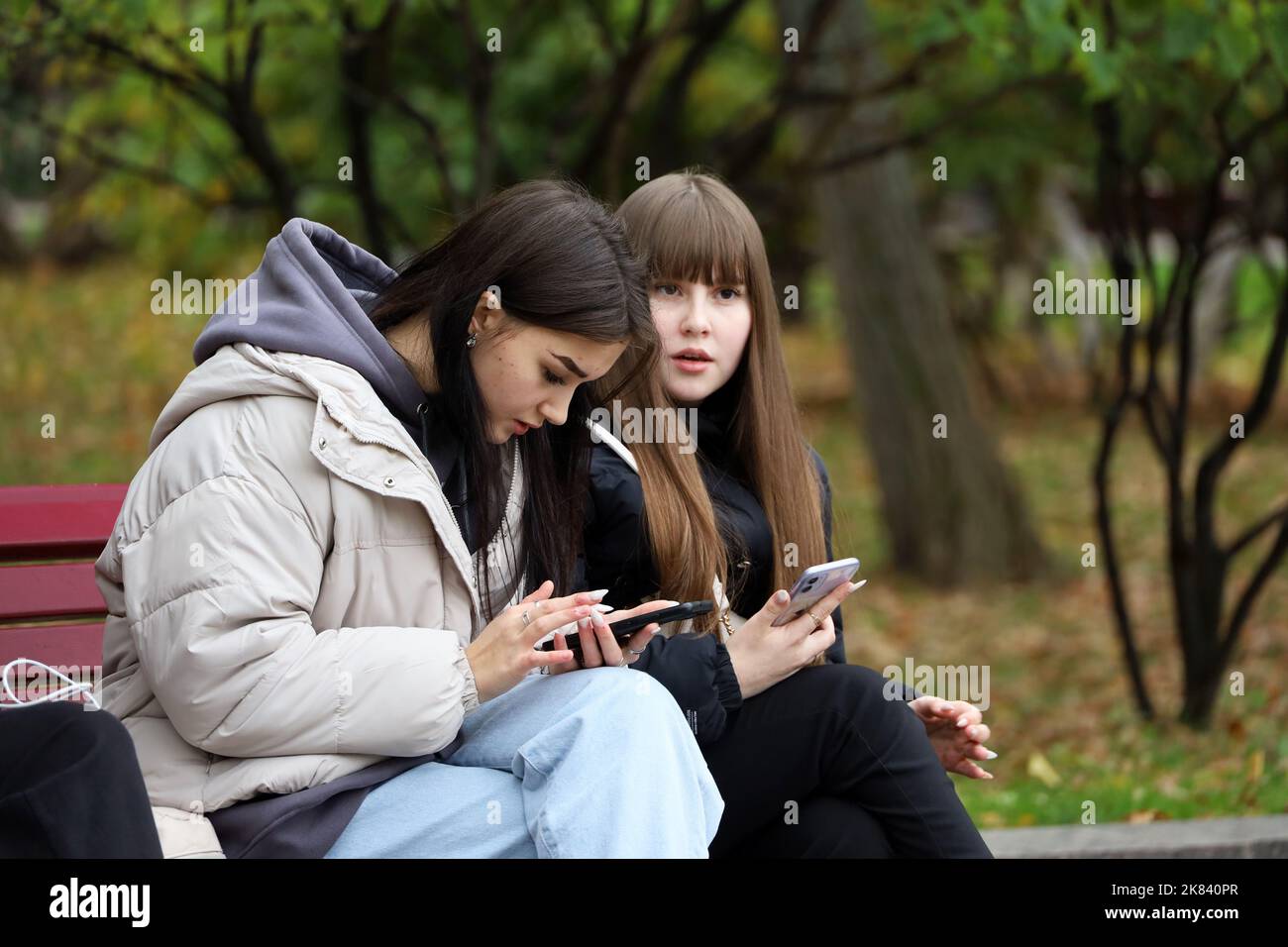 Due graziose ragazze in giacche sedute con smartphone su panca di legno nel parco autunnale. Concetto di dipendenza online, tempo libero in città Foto Stock