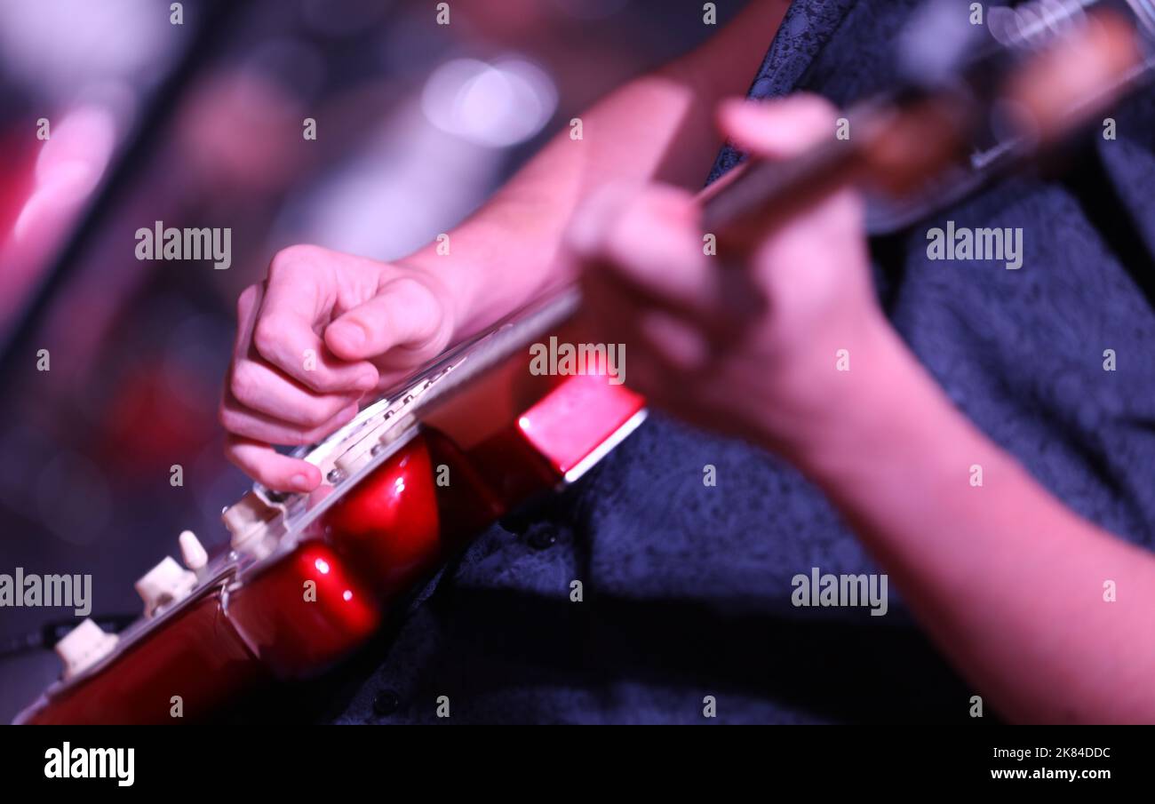 Primo piano della mano e delle dita che suonano musica su una chitarra elettrica rossa brillante. Il fret di strumming della mano con uno sfondo deliberatamente sfocato del riflettore Foto Stock